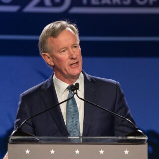 Admiral William McRaven, dressed in a suit and tie, speaks at a podium during an event, with a blue backdrop.